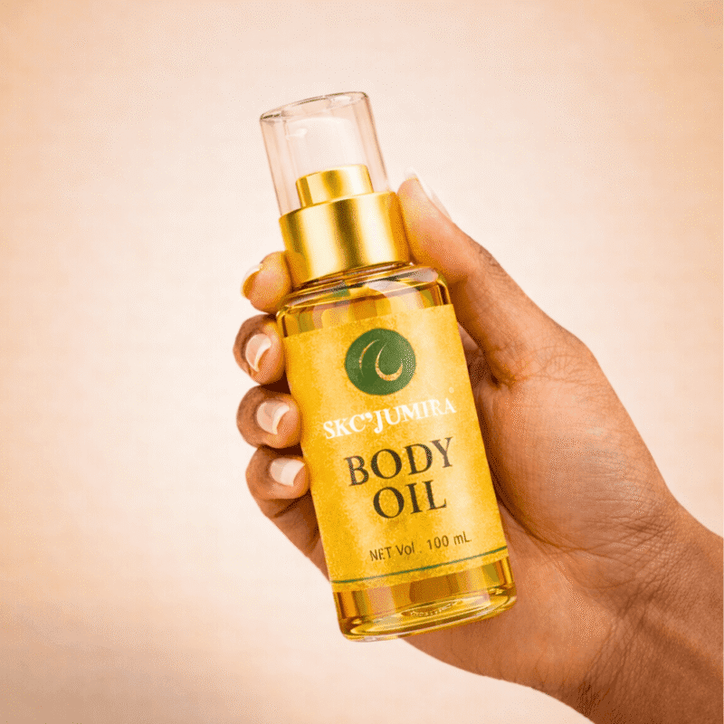 A hand holding a 100ml bottle of SKC’Jumira Body Oil with a gold pump, against a neutral background.